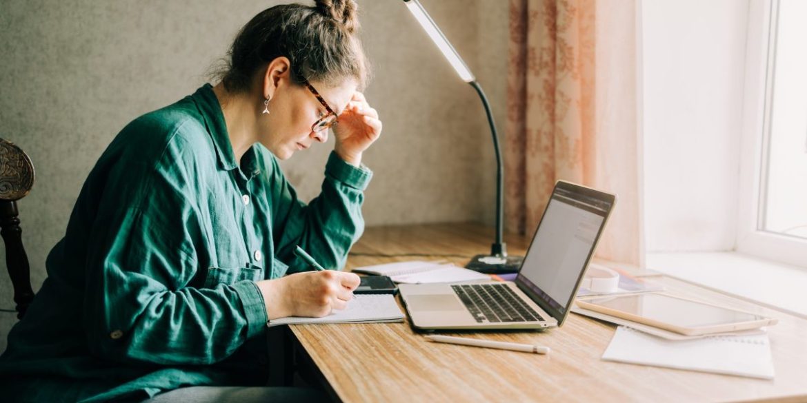 Stressed out woman looks at her notebook and computer while sitting at a desk.